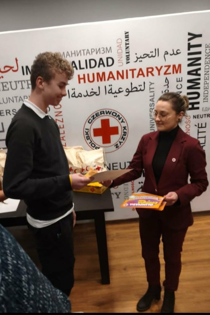 W sali konferencyjnej Pomorskiego Oddziału Okręgowego PCK na tle baneru PCK stoi uśmiechnięty uczeń, któremu organizatorka Olimpiady Zdrowia wręcza statuetkę jabłka, dyplom oraz nagrodę za zajęcie drugiego miejsca.