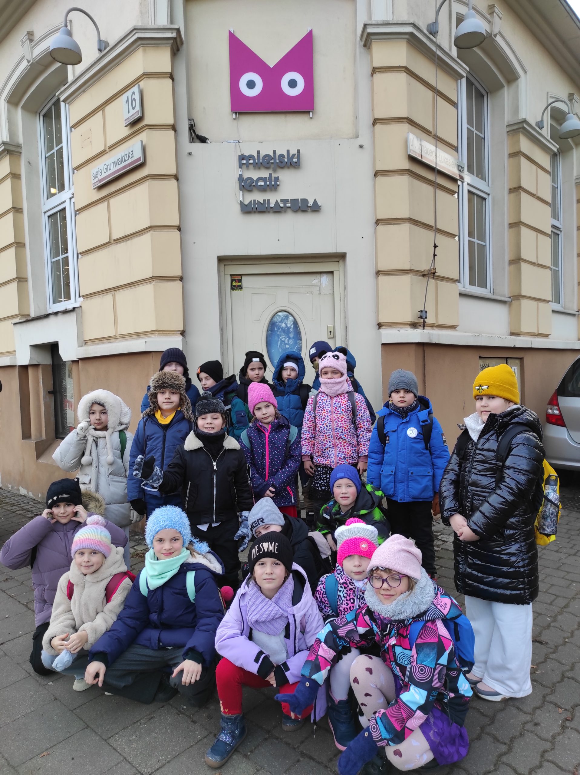 20 dzieci ustawionych w 2 rzędy, jeden stoi, drugi kuca. W tle fragment budynku, a na nim logo i nazwa obiektu : Miejski Teatr Miniatura.