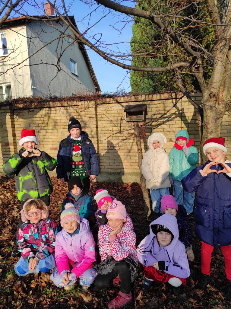 12 dzieci ustawionych w dwa rzędy. Jeden stoi, drugi siedzi. W tle karmnik dla ptaków, fragment drzewa oraz płotu.