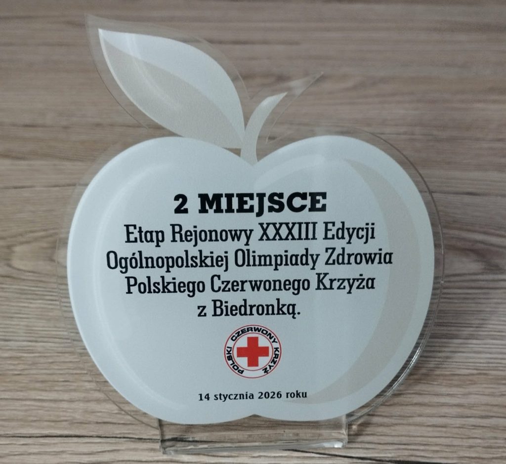 Na stole stoi statuetka jabłka z napisem 2 miejsce Etap Rejonowy XXXIII Edycji Ogólnopolskiej Olimpiady Zdrowia Polskiego Czewonego Krzyża z Biedronką oraz logo PCK.