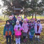 12 dzieci stoi w plenerze, przed karmnikiem dla ptaków. Jedna dziewczynka trzyma w dłoniach woreczek z pokarmem dla ptaków. Druga dotyka dłonią ziarnowe serce przyczepione do karmnika.