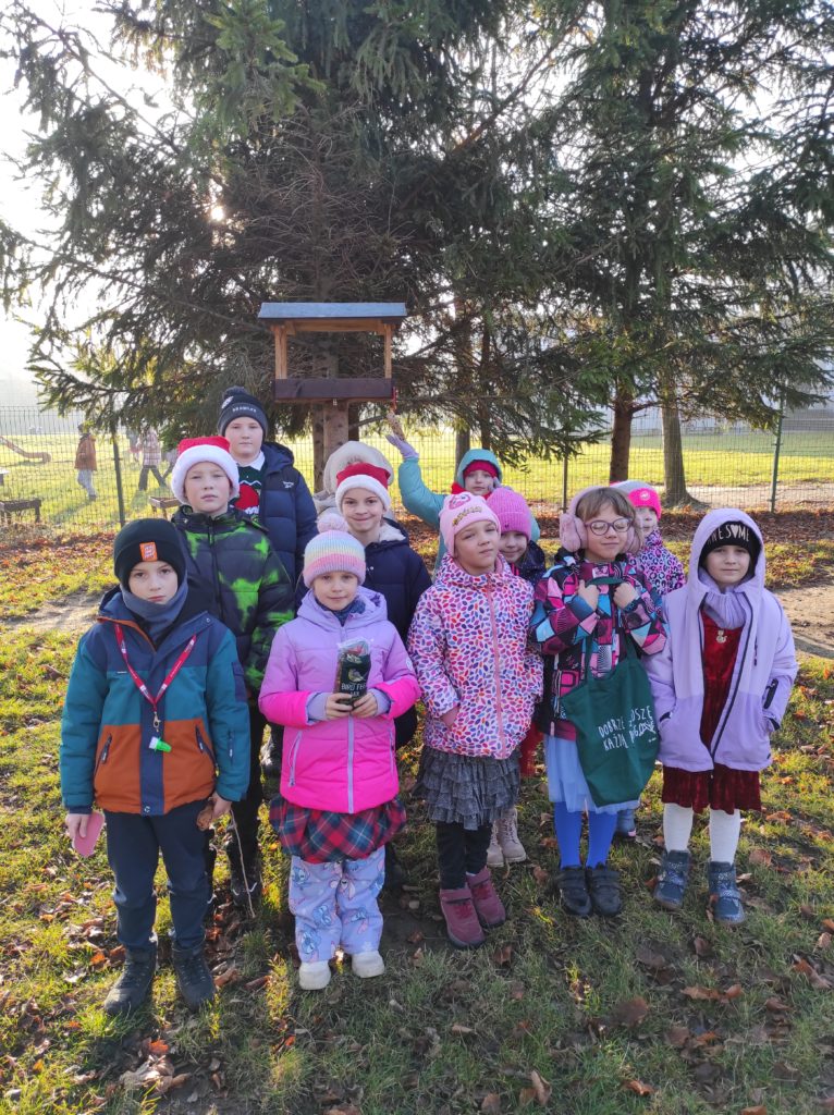 12 dzieci stoi w plenerze, przed karmnikiem dla ptaków. Jedna dziewczynka trzyma w dłoniach woreczek z pokarmem dla ptaków. Druga dotyka dłonią ziarnowe serce przyczepione do karmnika.