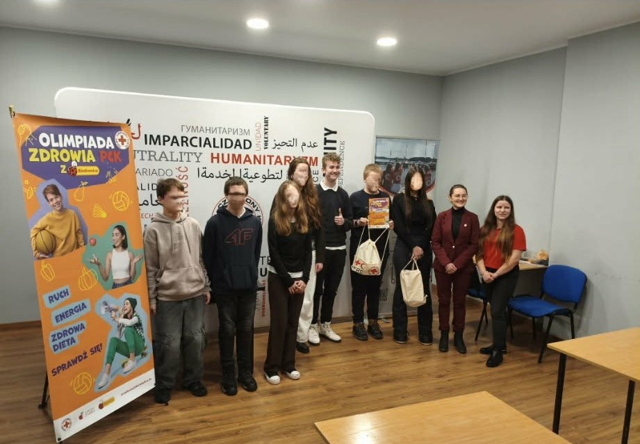 W sali konferencyjnej Pomorskiego Oddziału Okręgowego PCK na tle baneru PCK stoi 7 uczniów oraz uśmiechające się organizatorki Olimpiady Zdrowia.