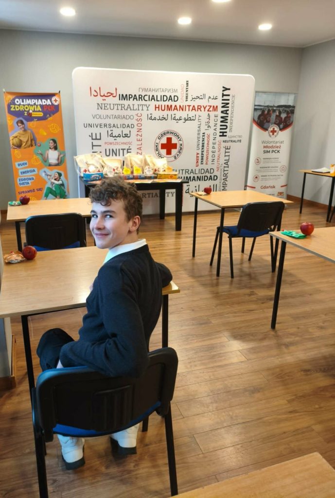 W sali konferencyjnej Pomorskiego Oddziału Okręgowego PCK na tle baneru PCK oraz nagród siedzi uczeń przy stoliku.