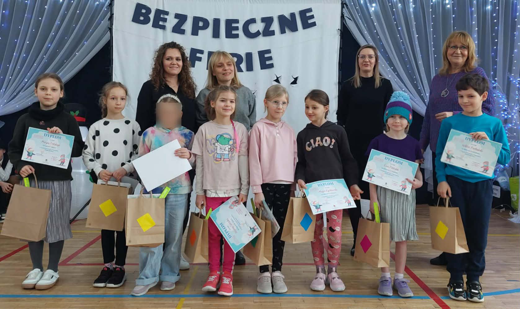 Grupa uczniów klas I–III stoi na sali gimnastycznej podczas podsumowania konkursu plastycznego „Bezpieczne Ferie”. Dzieci trzymają dyplomy i torby z nagrodami. Za nimi stoją nauczyciele, a w tle widoczny jest baner z nazwą konkursu.