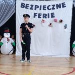 Uczeń przebrany za policjanta stoi na szkolnej scenie i trzyma mikrofon. W tle dekoracja z napisem „Bezpieczne ferie”, zimowe ozdoby oraz dmuchane bałwanki.