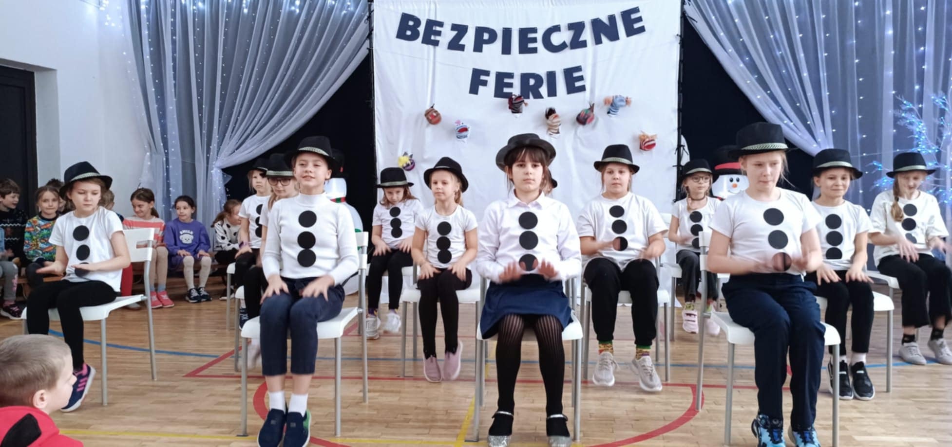 Grupa dzieci siedzi na krzesłach na sali gimnastycznej i wykonuje układ taneczny. Uczniowie mają stroje stylizowane na bałwanki, w tle scena z napisem „Bezpieczne ferie”.