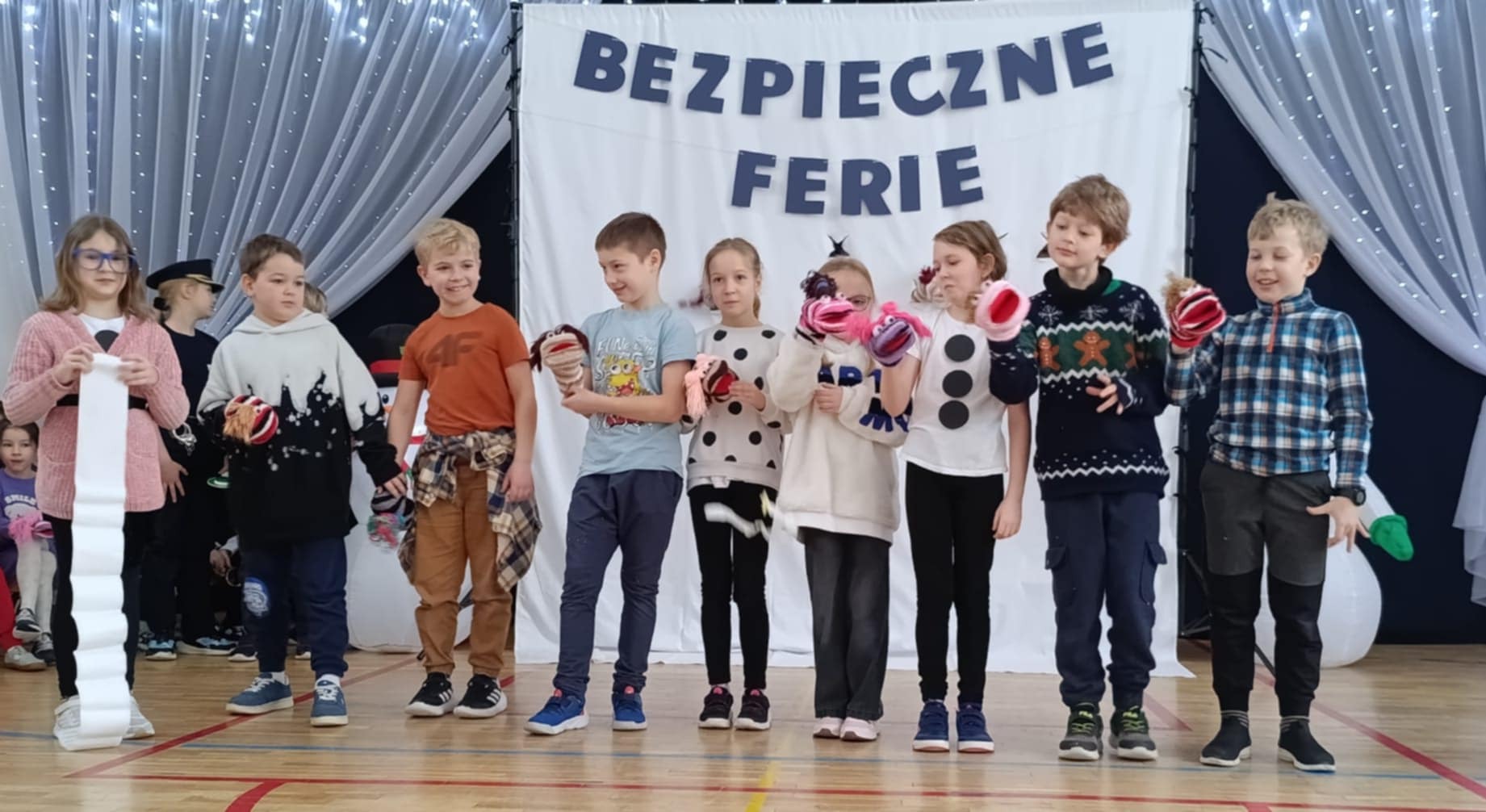 Dzieci ustawione w rzędzie stoją na szkolnej scenie. Mają w rękach pacynki do przedstawienia, a za nimi wisi dekoracja z napisem „Bezpieczne ferie”.