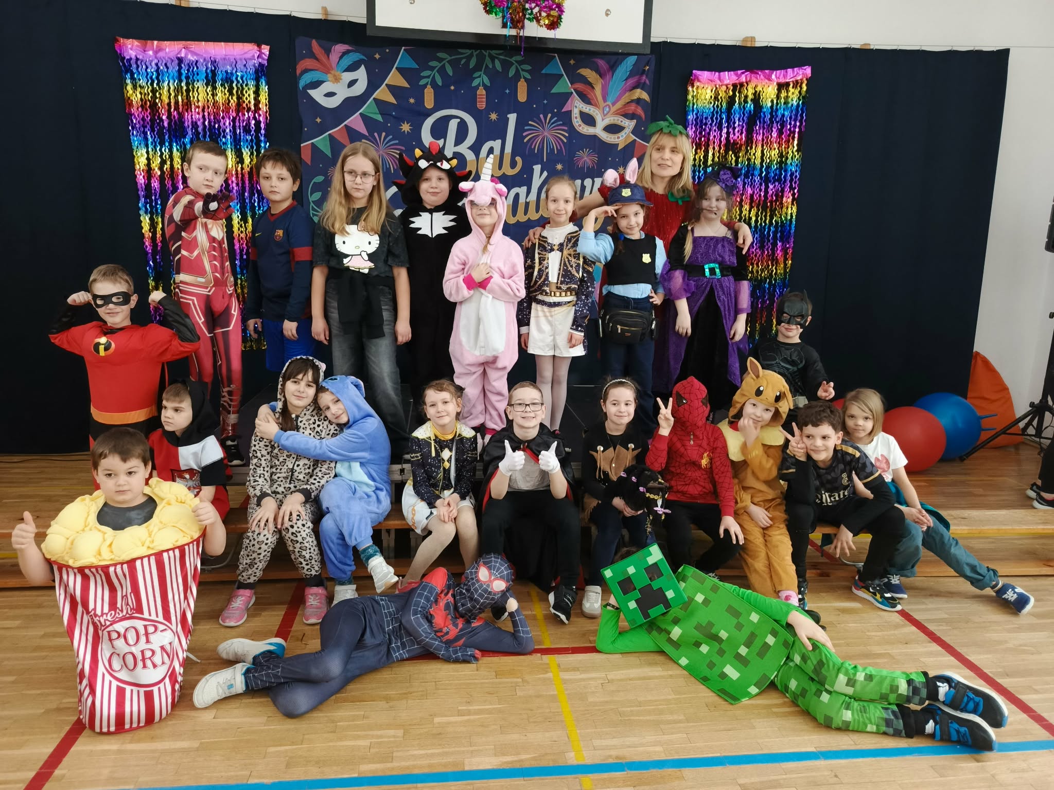 przesyłam 2 zdjęcia z opisem: Grupowe zdjęcie dzieci podczas balu karnawałowego w szkolnej sali gimnastycznej. Uczniowie stoją i siedzą na podwyższeniu przed dekoracyjną ścianką z napisem „Bal karnawałowy”, kolorowymi maskami i tęczowymi kurtynami. Dzieci mają na sobie różnorodne stroje. W pierwszym rzędzie część uczniów siedzi na podłodze, dwóch chłopców leży w przebraniach superbohatera i postaci z gry komputerowej.