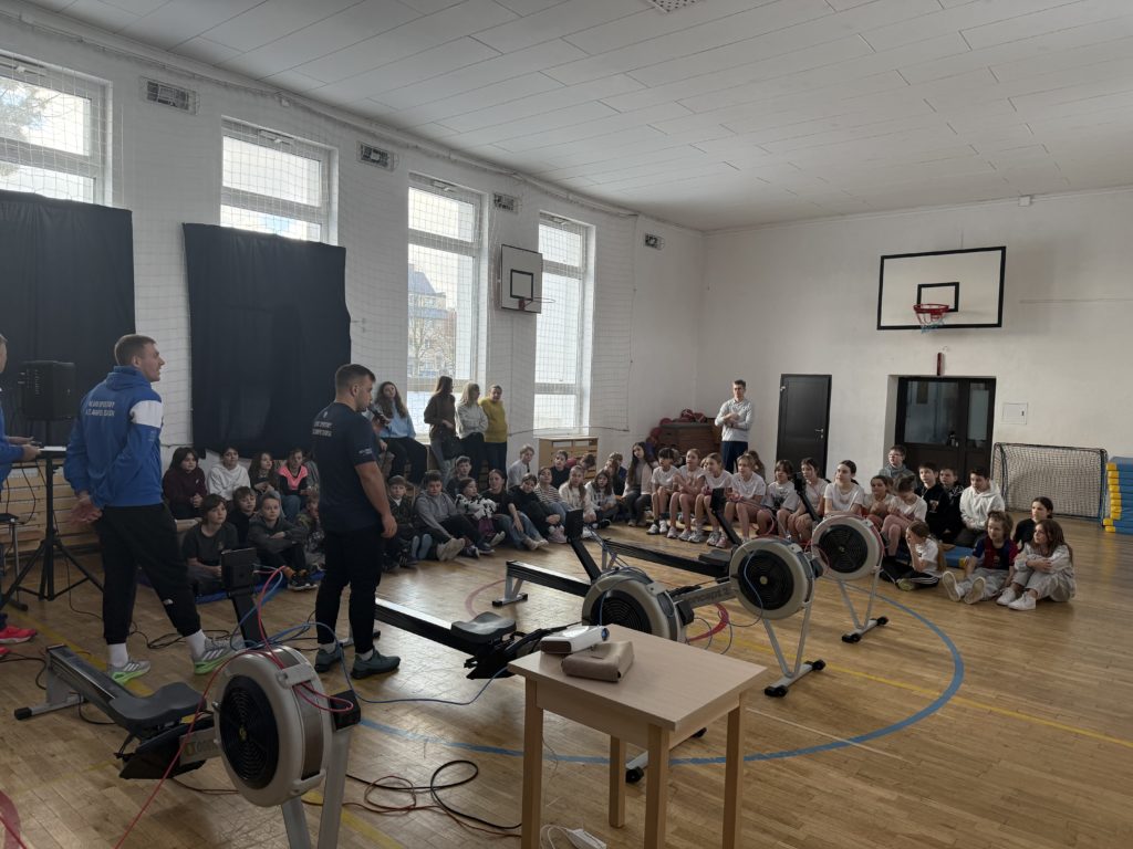 W sali gimnastycznej dwaj mężczyźni prowadzą spotkanie dla grupy uczniów siedzących na pod-łodze i ławkach pod ścianą. Przed nimi ustawione są ergometry wioślarskie podłączone do monito-rów. W tle widać kosze do koszykówki, wysokie okna oraz nauczycieli stojących przy ścianie.