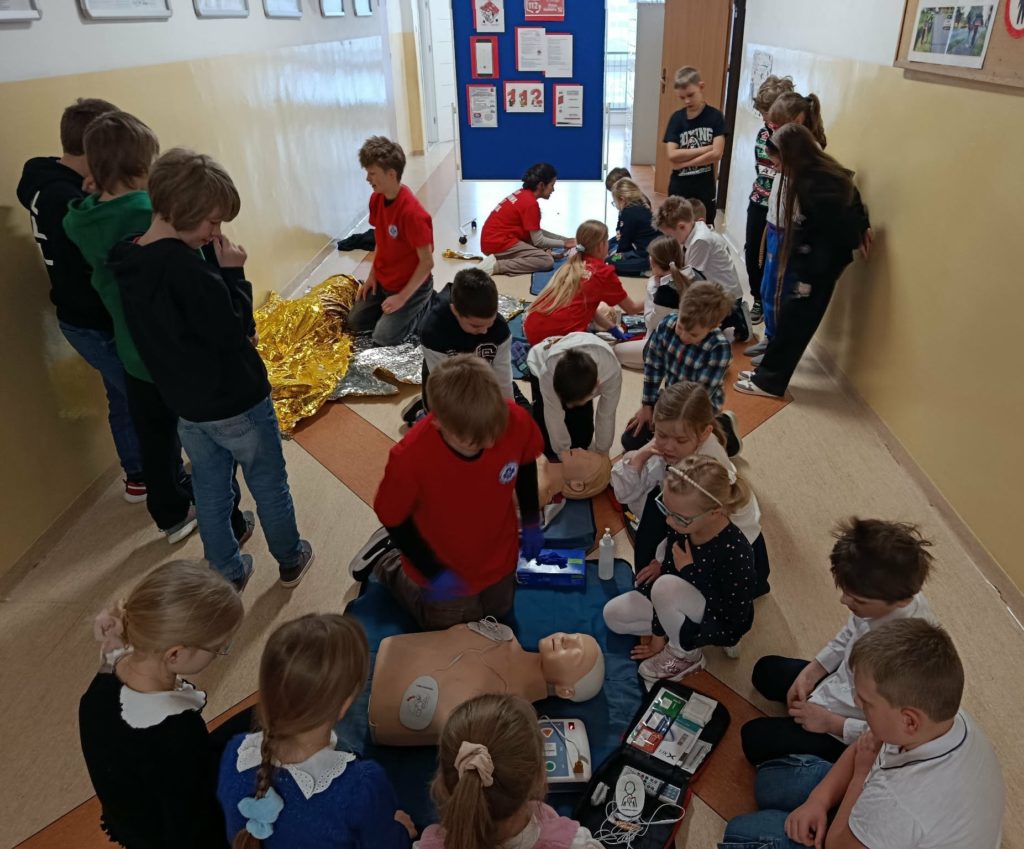 Na szkolnym korytarzu na tle tablicy z plakatem na temat numeru alarmowego 112 stoi lub kuca liczna grupa uczniów. Na pierwszym planie uczeń Koła PP HOPR przy fantomie z elektrodami AED. W koło przyglądający się uczniowie.