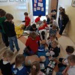 Na szkolnym korytarzu na tle tablicy z plakatem na temat numeru alarmowego 112 stoi lub kuca liczna grupa uczniów. Na pierwszym planie uczeń Koła PP HOPR przy fantomie z elektrodami AED. W koło przyglądający się uczniowie.