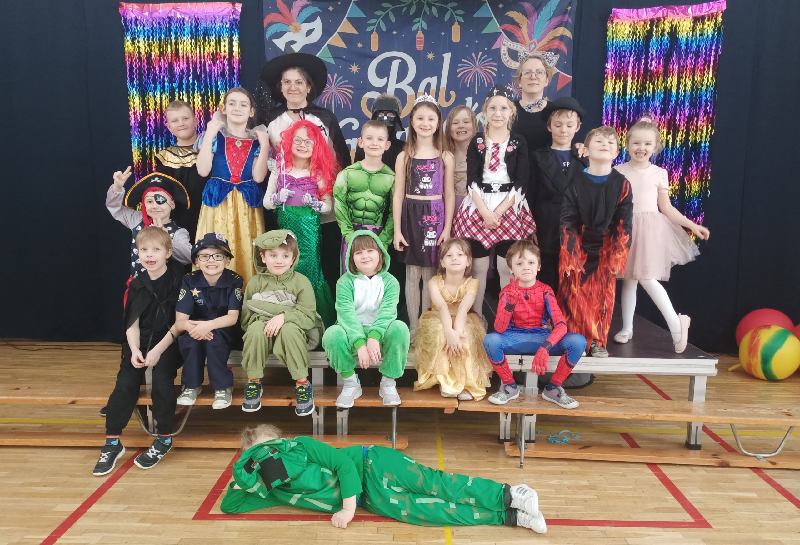 Zdjęcie grupowe dzieci i nauczycielki podczas balu karnawałowego. W tle widać granatową kotarę z napisem „Bal Karnawałowy”, kolorowe maski oraz błyszczące, tęczowe girlandy. Dzieci są przebrane w różnorodne kostiumy, m.in. Superbohaterów, księżniczki, zwierzęta i postacie z bajek.