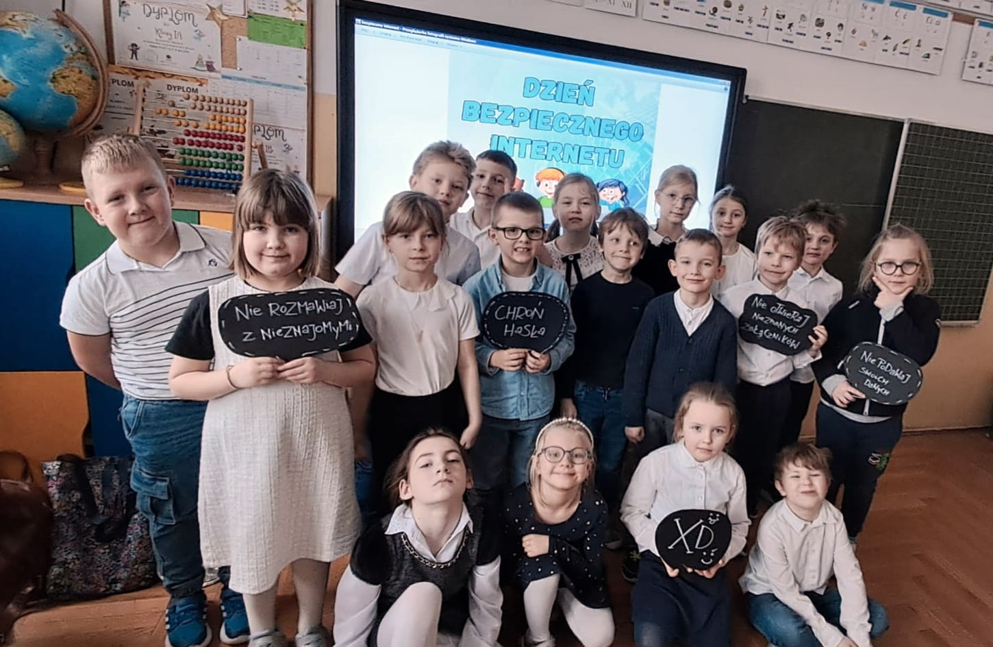 Grupa uśmiechniętych dzieci w wieku wczesnoszkolnym stoi w klasie na tle tablicy interaktywnej z napisem „Dzień Bezpiecznego Internetu”. Niektóre dzieci trzymają czarne tabliczki w kształcie chmurek z hasłami w języku polskim, m.in.: „Nie rozmawiaj z nieznajomymi”, „Chroń hasło” oraz „Nie podawaj swoich danych”.