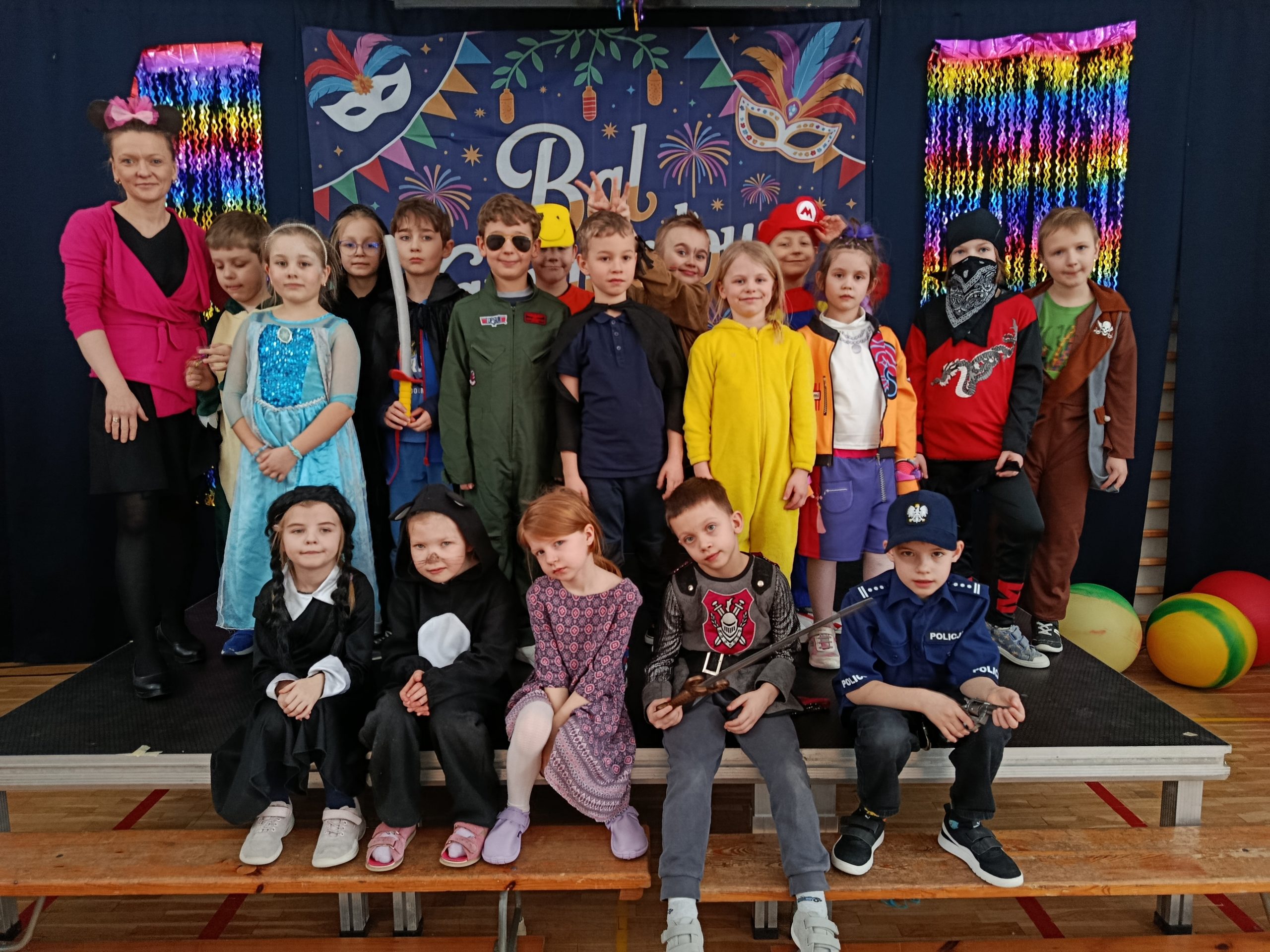 Zdjęcie grupowe dzieci i nauczycielki podczas balu karnawałowego. W tle widać granatową kotarę z napisem „Bal Karnawałowy”, kolorowe maski oraz błyszczące, tęczowe girlandy. Dzieci są przebrane w różnorodne kostiumy, m.in. Superbohaterów, księżniczki, zwierzęta i postacie z bajek.