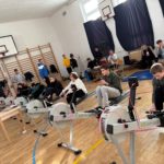 W sali gimnastycznej kilku chłopców siedzi na ergometrach wioślarskich ustawionych w rzędzie i przygotowuje się do ćwiczeń. Urządzenia są podłączone przewodami do monitorów. W tle stoją inni uczniowie oraz dorośli obserwujący wydarzenie. Na ścianach widać drabinki gimnastyczne i kosze do koszykówki.