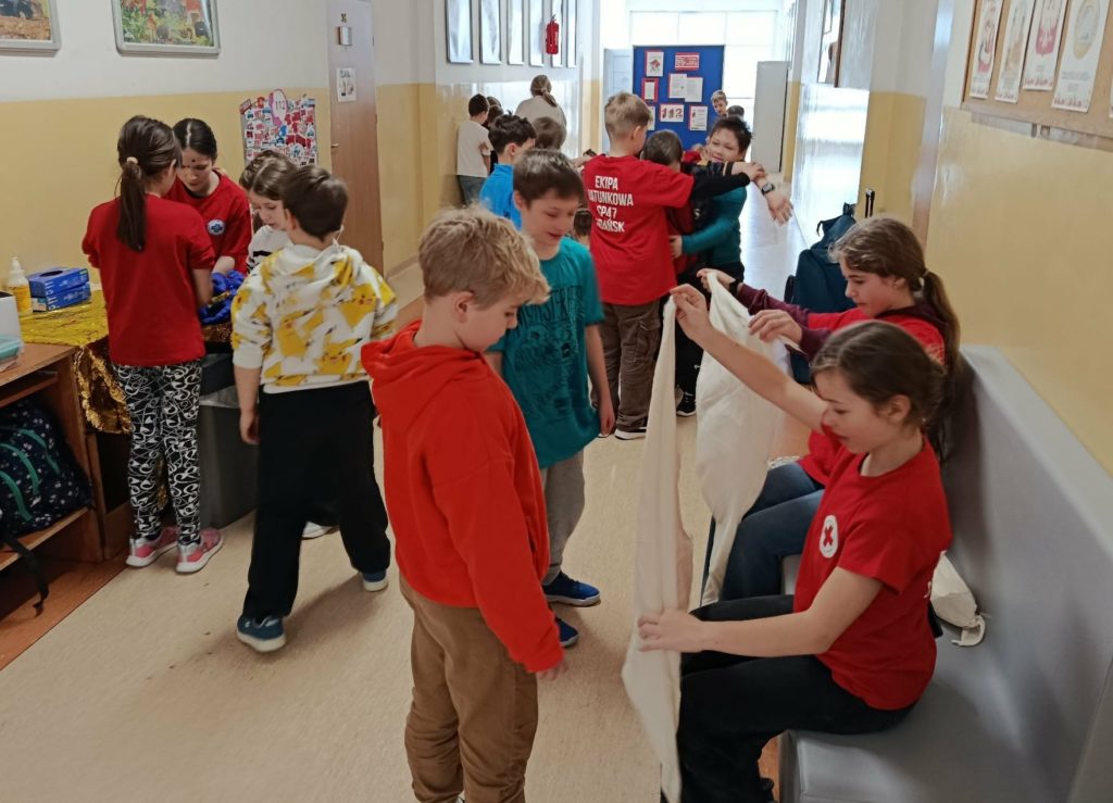 Na szkolnym korytarzu dwie uczennice Koła PP HOPR demonstrujące chłopcom zakładanie temblaka i uczennica demonstrująca bezpieczne zdejmowanie rękawiczek. W tle uczeń uczący odkrztuszania na kamizelce Act Fast.