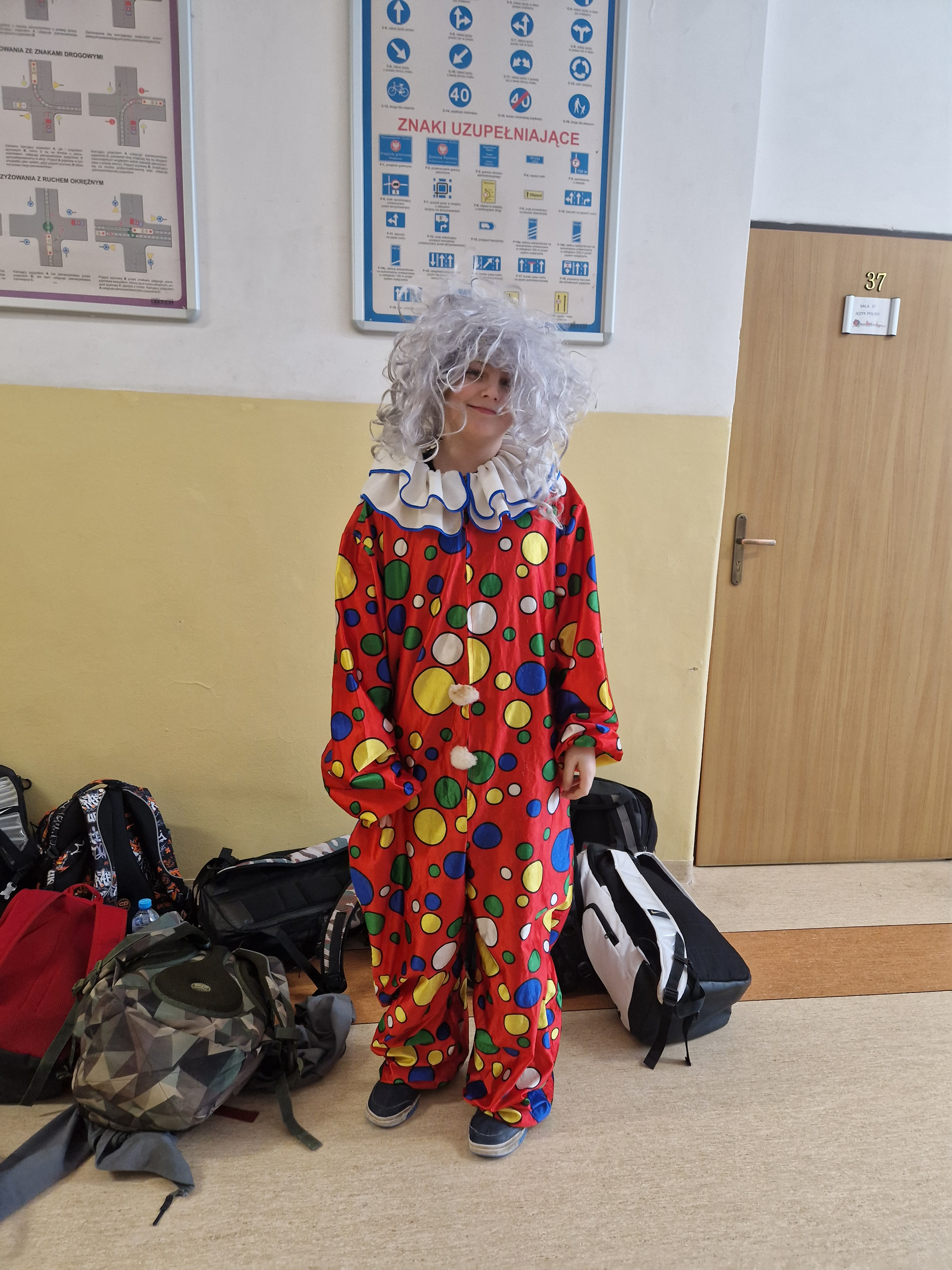 Dziecko przebrane za klauna stoi w szkolnym korytarzu. Ma na sobie czerwony kostium w kolorowe kropki, białą perukę i szeroki kołnierz. Obok leżą plecaki, a na ścianie wiszą tablice z przepisami ruchu drogowego.