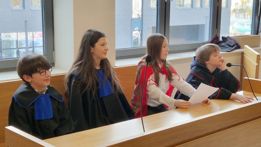 Czworo dzieci siedzi przy ławie w sali sądowej; troje w togach, jedna dziewczynka w stroju ludowym trzyma kartkę, przed nimi stoją mikrofony.