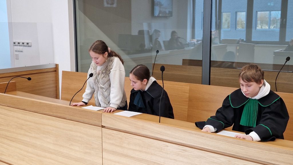 Troje dzieci siedzi przy ławie w sali sądowej patrzą na kartki; dwoje ma na sobie togi, przed nimi stoją mikrofony.