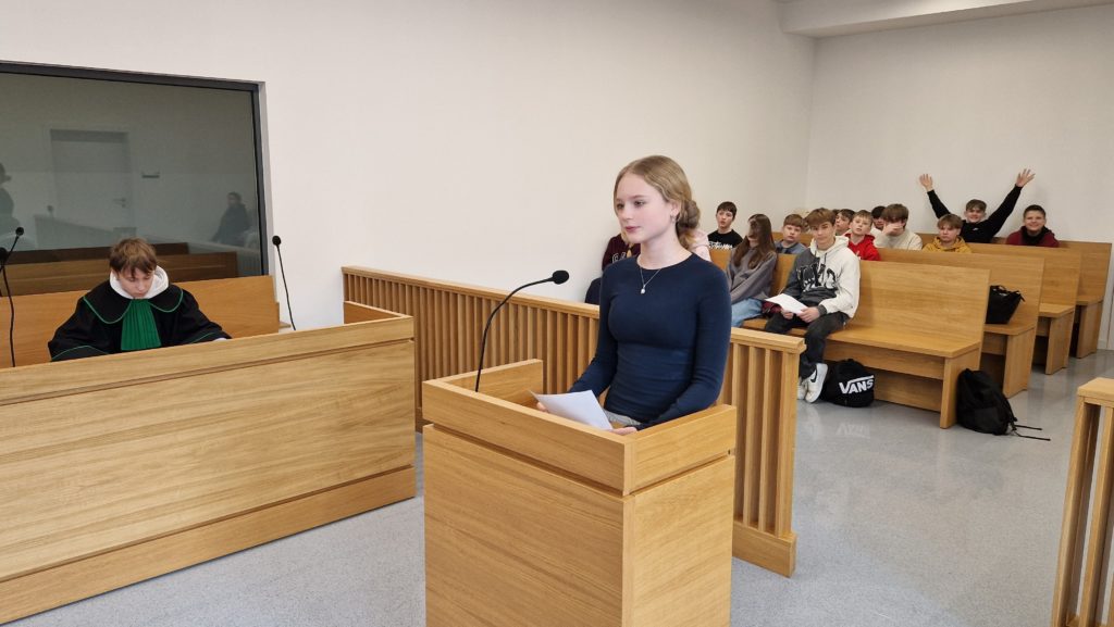 Nastolatka stoi przy mównicy w sali sądowej i czyta z kartki, w tle siedzi sędzia oraz grupa uczniów na ławkach.