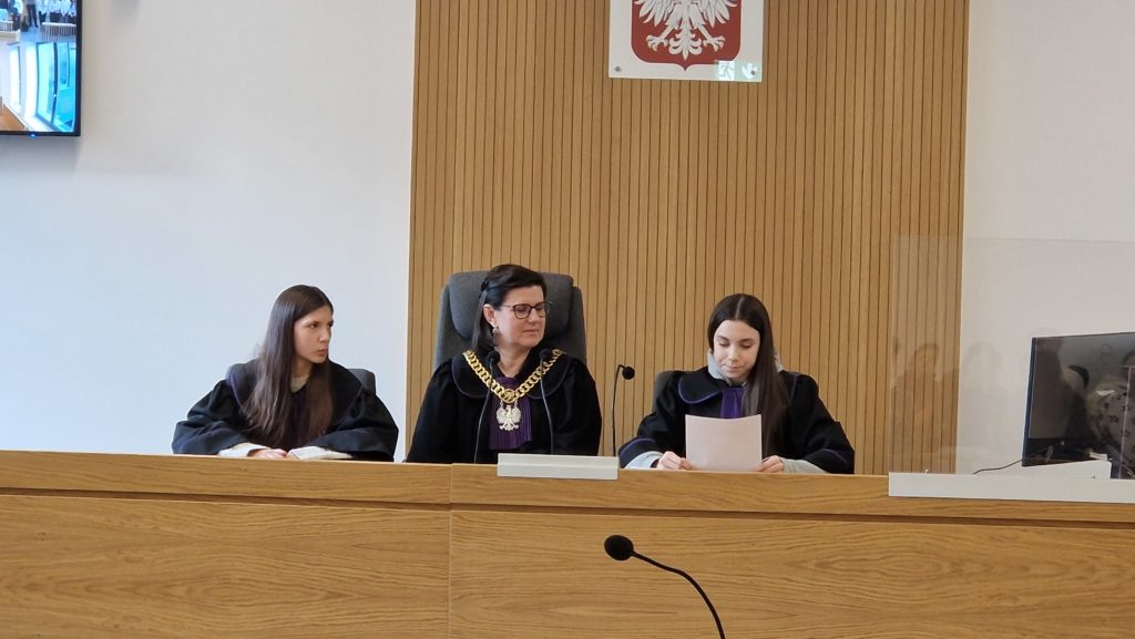Trzy kobiety w togach siedzą przy stole sędziowskim w sali sądowej; pośrodku sędzia z łańcuchem z godłem, w tle godło Polski.