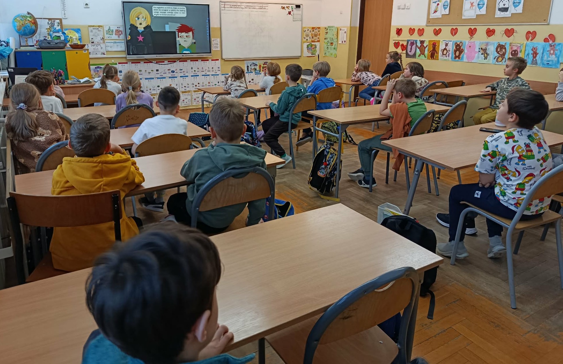 Widok z tyłu klasy na uczniów siedzących w ławkach. Dzieci patrzą na tablicę interaktywną, na której wyświetlany jest film edukacyjny z postaciami z kreskówek. Sala lekcyjna jest kolorowa, na ścianach wiszą prace plastyczne uczniów przedstawiające postacie i zwierzęta.