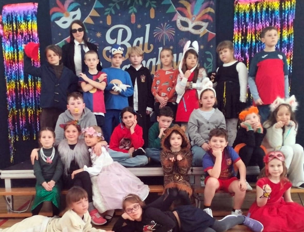 Zdjęcie grupowe dzieci i nauczycielki podczas balu karnawałowego. W tle widać granatową kotarę z napisem „Bal Karnawałowy”, kolorowe maski oraz błyszczące, tęczowe girlandy. Dzieci są przebrane w różnorodne kostiumy, m.in. Superbohaterów, księżniczki, zwierzęta i postacie z bajek.