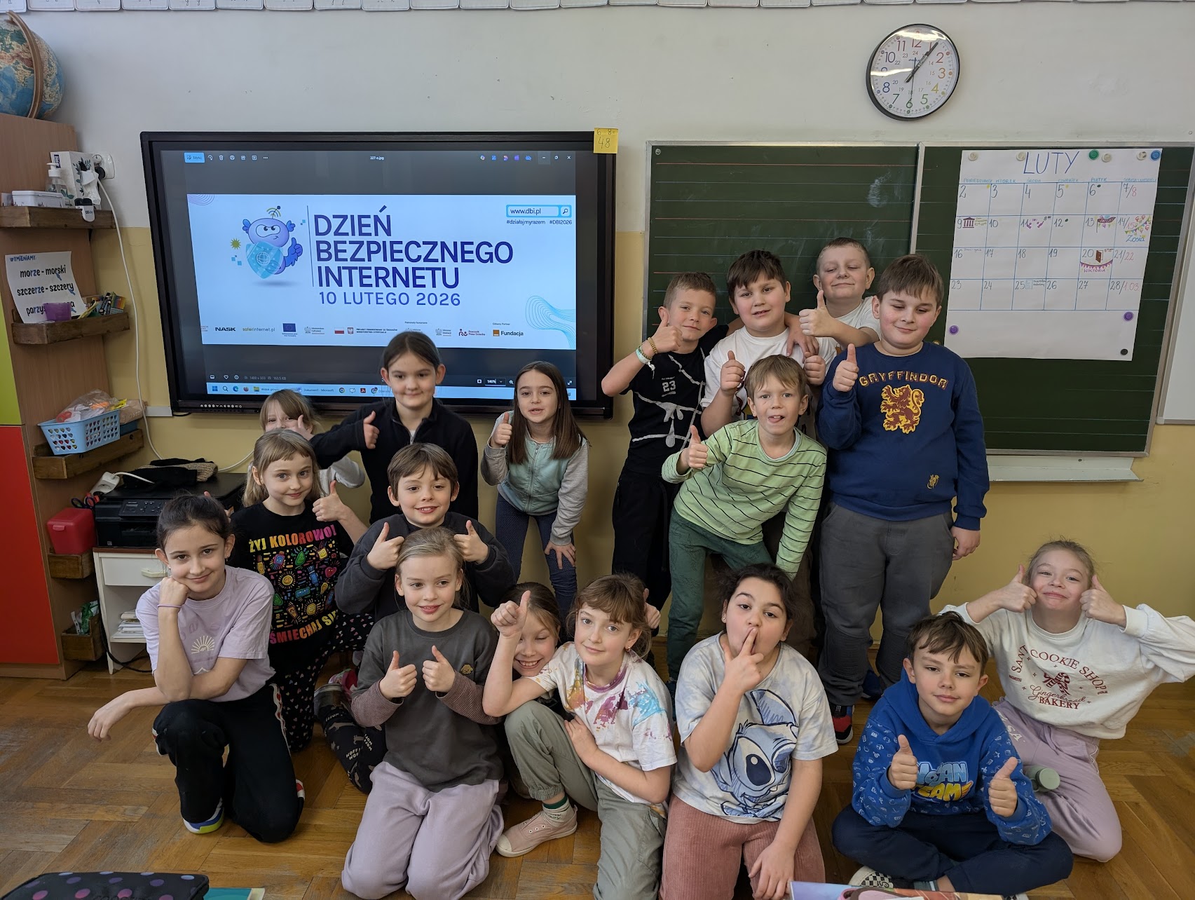 Grupa dzieci pozuje do wspólnego zdjęcia w klasie, wykonując gest kciuka w górę. W tle znajduje się ekran z oficjalną grafiką „Dzień Bezpiecznego Internetu – 10 lutego 2026” oraz zielona tablica z zawieszonym kalendarzem na luty. Atmosfera jest radosna i entuzjastyczna.