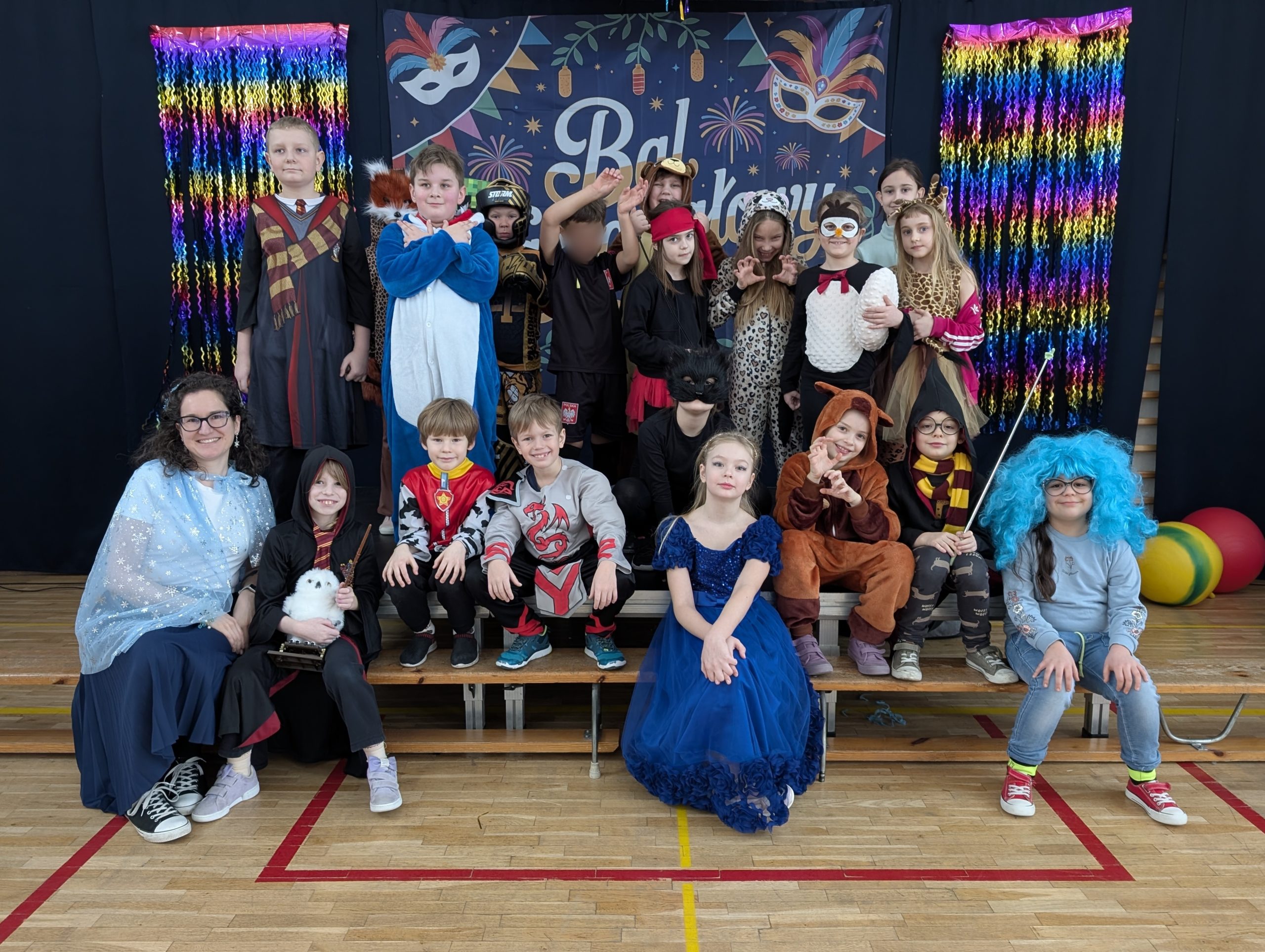 Zdjęcie grupowe dzieci i nauczycielki podczas balu karnawałowego. W tle widać granatową kotarę z napisem „Bal Karnawałowy”, kolorowe maski oraz błyszczące, tęczowe girlandy. Dzieci są przebrane w różnorodne kostiumy, m.in. Superbohaterów, księżniczki, zwierzęta i postacie z bajek.