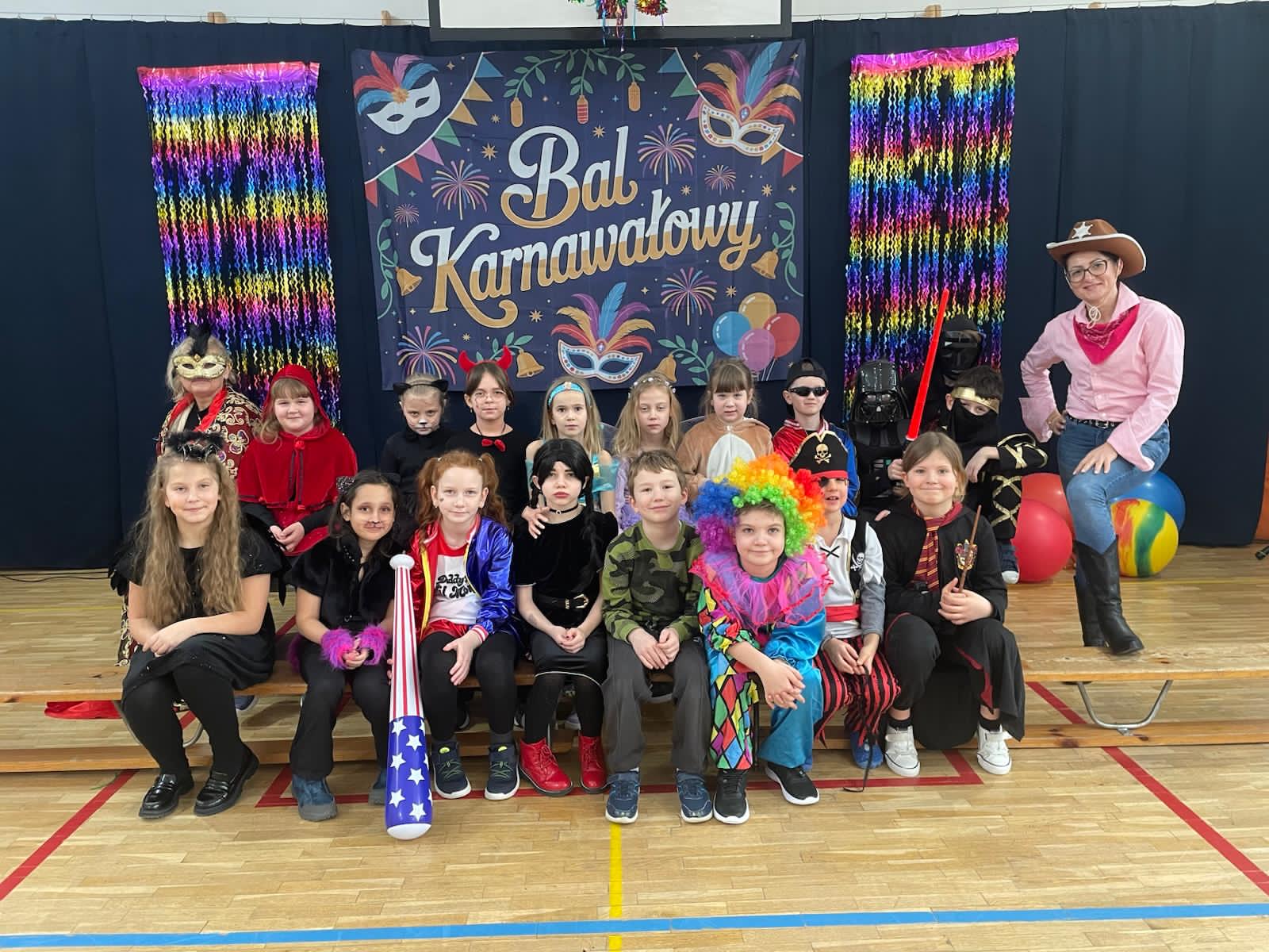Zdjęcie grupowe dzieci i nauczycielki podczas balu karnawałowego. W tle widać granatową kotarę z napisem „Bal Karnawałowy”, kolorowe maski oraz błyszczące, tęczowe girlandy. Dzieci są przebrane w różnorodne kostiumy, m.in. Superbohaterów, księżniczki, zwierzęta i postacie z bajek.