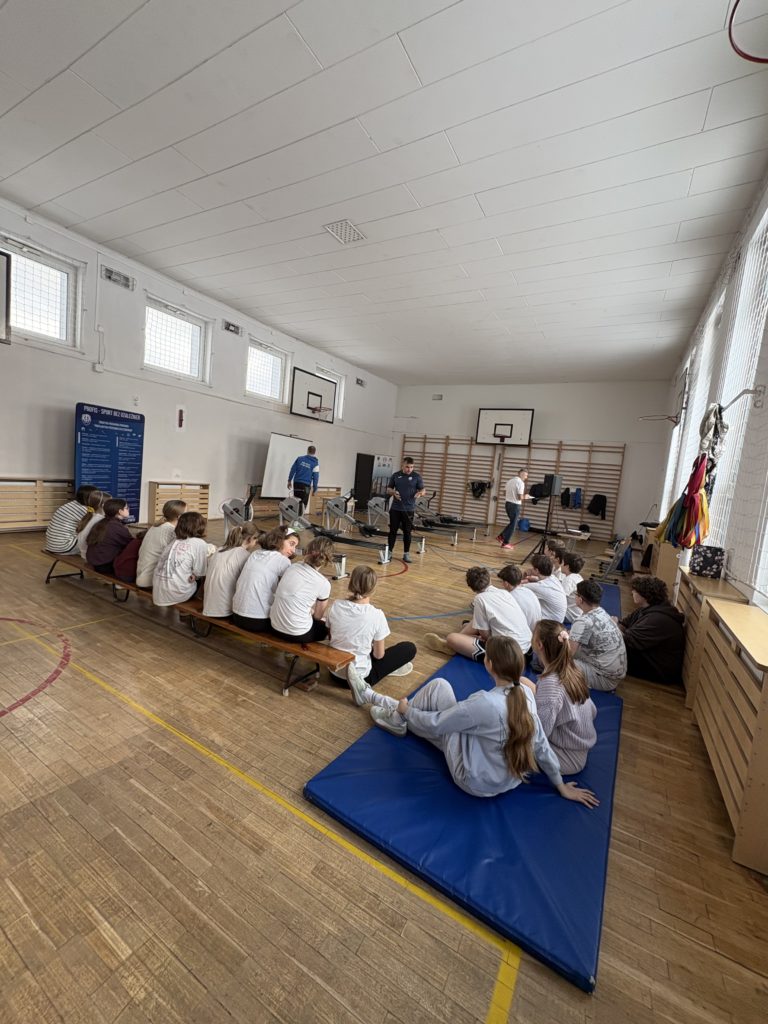 W sali gimnastycznej grupa uczniów siedzi na ławkach i materacu, słuchając prowadzących stoją-cych przy ergometrach wioślarskich ustawionych na środku sali. W tle widać drabinki gimna-styczne, kosze do koszykówki oraz sprzęt sportowy.