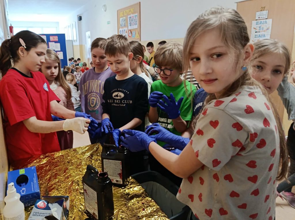 Na szkolnym korytarzu uczennica demonstrująca bezpieczne zakładanie rękawiczek jednorazowych. W koło uczniowie zdejmujący zabrudzone rękawiczki.