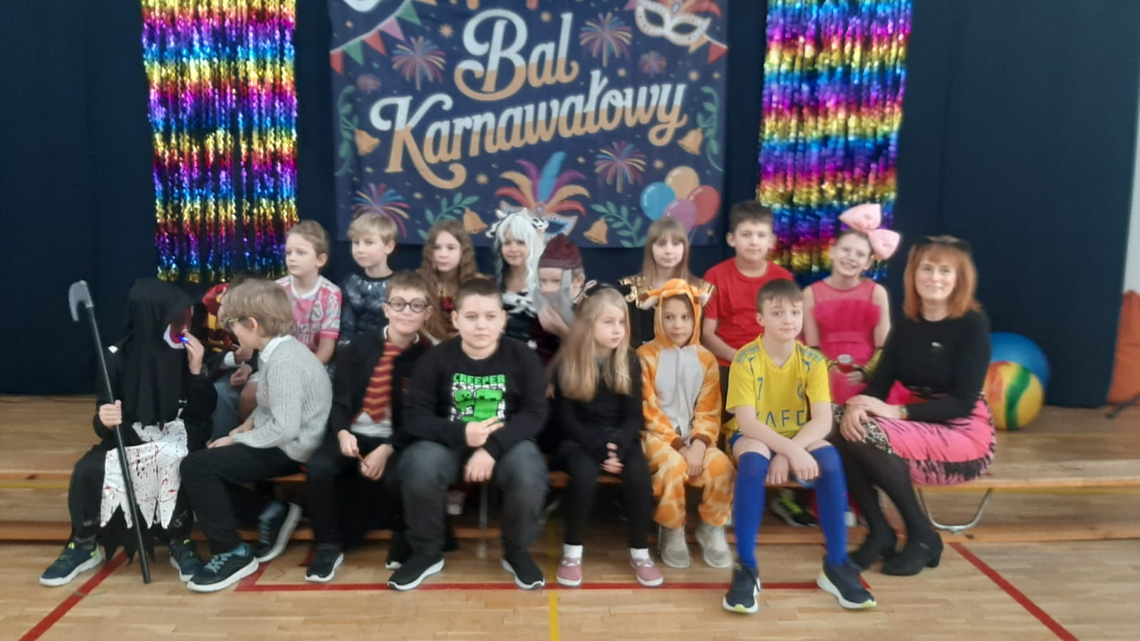 Zdjęcie grupowe dzieci i nauczycielki podczas balu karnawałowego. W tle widać granatową kotarę z napisem „Bal Karnawałowy”, kolorowe maski oraz błyszczące, tęczowe girlandy. Dzieci są przebrane w różnorodne kostiumy, m.in. Superbohaterów, księżniczki, zwierzęta i postacie z bajek.