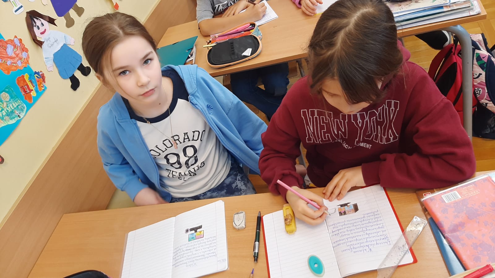 Dwie dziewczynki siedzą przy szkolnej ławce i pracują w zeszytach. Jedna z nich koloruje rysunek przedstawiający komputer, druga patrzy w stronę obiektywu. Na ławce leżą przybory szkolne: gumka, temperówka i długopisy. W tle na ścianie widoczne są wycinanki wykonane przez dzieci.