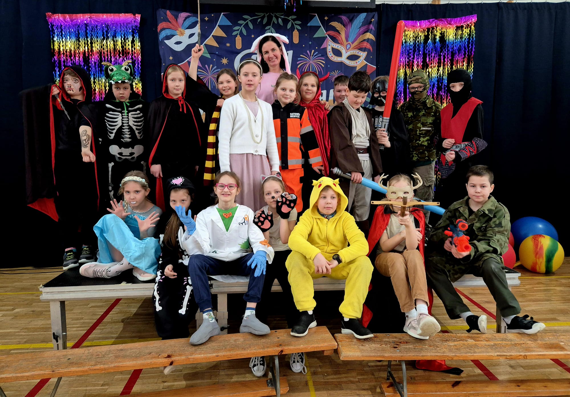 Zdjęcie grupowe dzieci i nauczycielki podczas balu karnawałowego. W tle widać granatową kotarę z napisem „Bal Karnawałowy”, kolorowe maski oraz błyszczące, tęczowe girlandy. Dzieci są przebrane w różnorodne kostiumy, m.in. Superbohaterów, księżniczki, zwierzęta i postacie z bajek.