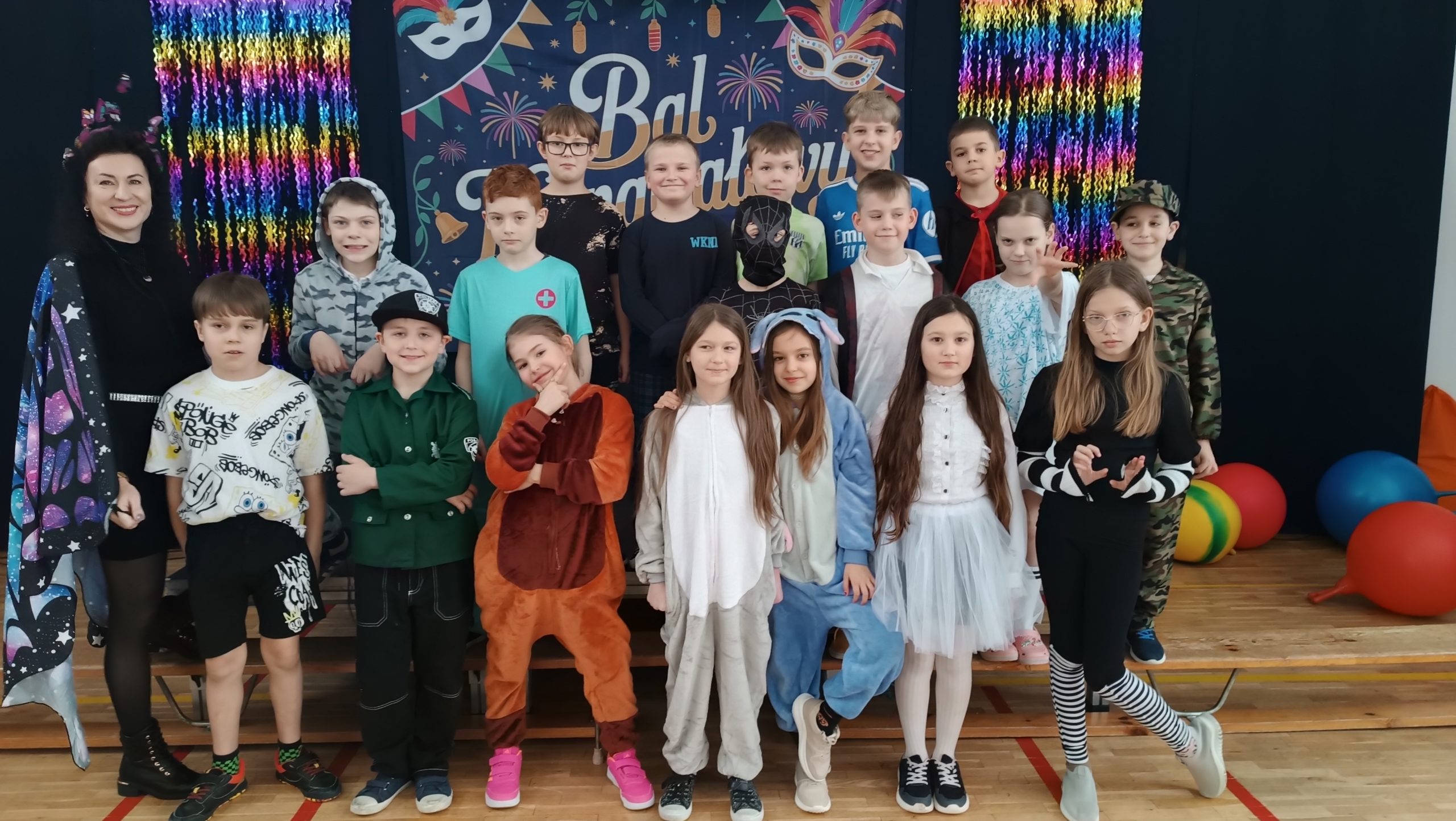 Zdjęcie grupowe dzieci i nauczycielki podczas balu karnawałowego. W tle widać granatową kotarę z napisem „Bal Karnawałowy”, kolorowe maski oraz błyszczące, tęczowe girlandy. Dzieci są przebrane w różnorodne kostiumy, m.in. Superbohaterów, księżniczki, zwierzęta i postacie z bajek.