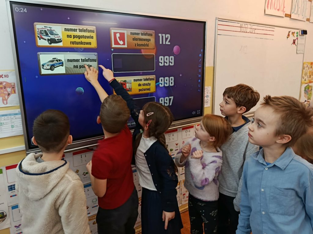 W sali lekcyjnej na tablicy multimedialnej uczniowie klasy pierwszej grający w grę utrwalającą numery alarmowe.