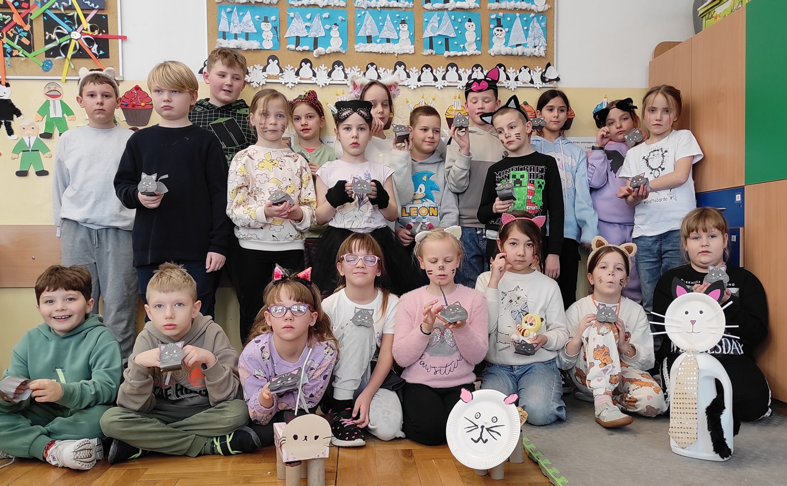 Dzieci ustawione w trzech rzędach, dwa stoją, jeden przedni siedzi. Niektóre z nich mają kocie akcenty w ubiorze. Każdy trzyma w dłoniach małego kotka z papieru. Przed dziećmi są ustawione trzy koty wykonane z surowców wtórnych. W tle elementy sali.