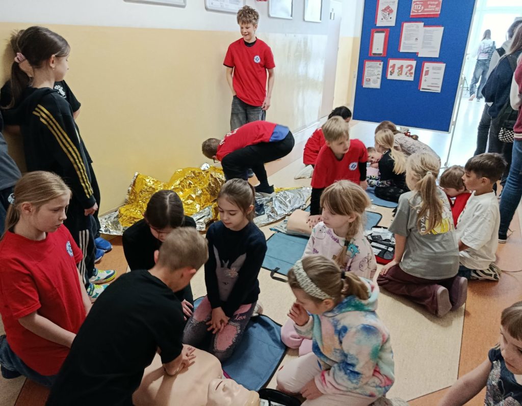 Na szkolnym korytarzu na tle tablicy z plakatem na temat numeru alarmowego 112 uczniowie Koła PP HOPR demonstrujący RKO na fantomach, uczniowie pokazujący pozycję bezpieczną oraz liczna grupa przyglądających się uczniów klas młodszych.