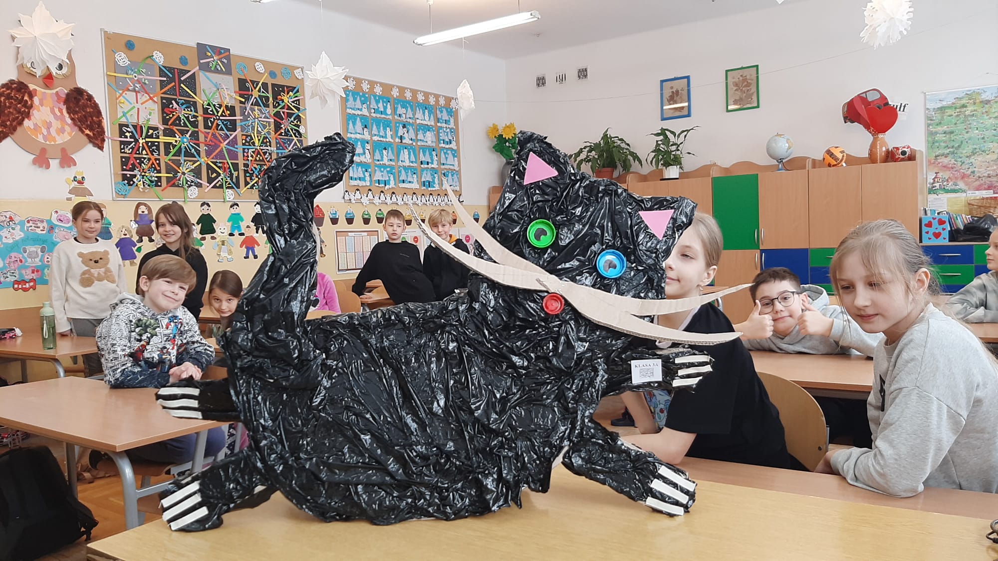 a pierwszym planie duży kot wykonany z surowców wtórnych oraz trzy ławki, a w nich 5 dzieci. Jeden chłopiec klęczy z lewej strony, przy ławce. W tle elementy sali.