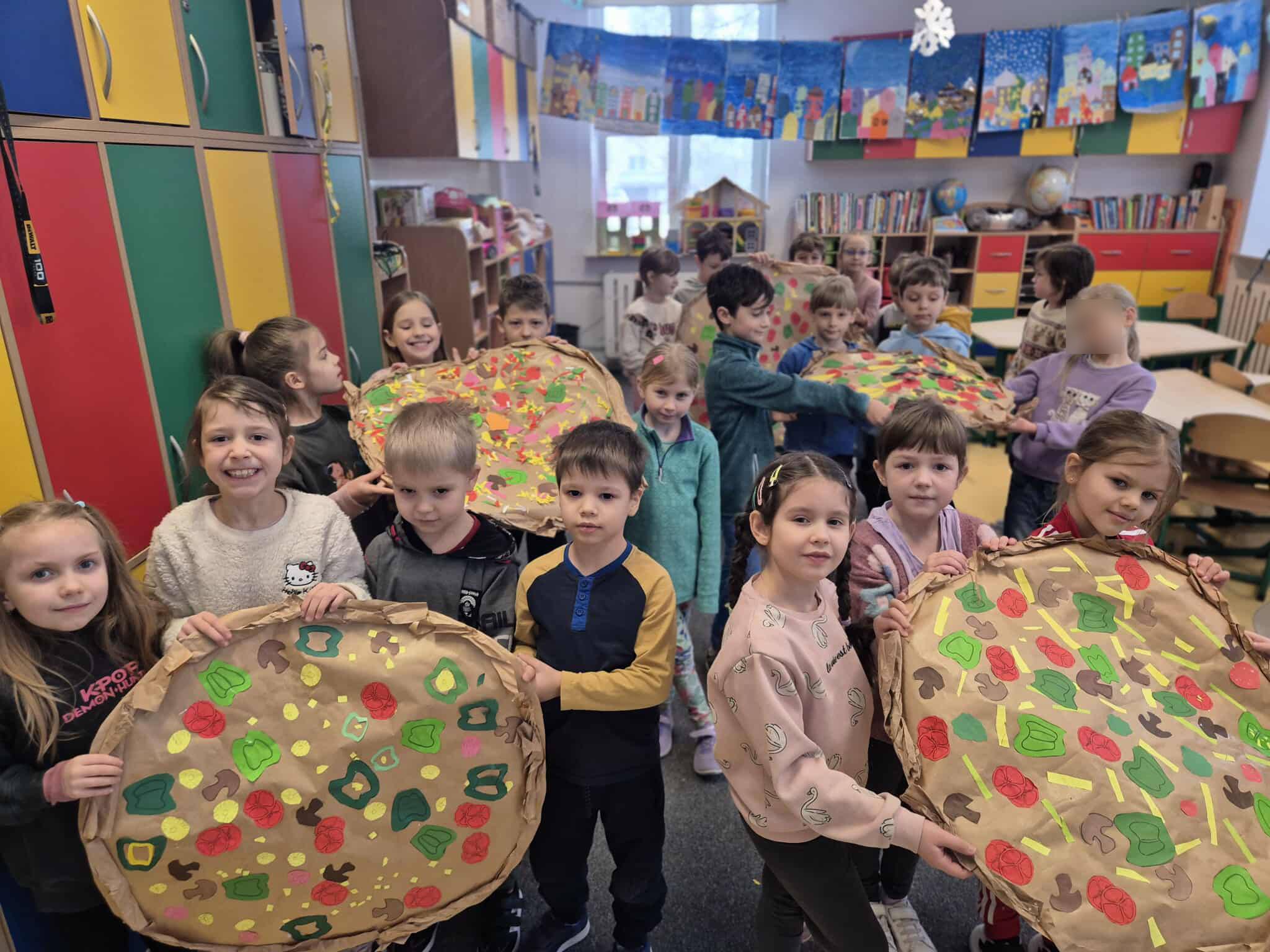 Grupa dzieci stoi w sali i trzyma kilka dużych, okrągłych prac plastycznych przedstawiających pizze z papieru. Na każdej pizzy widać kolorowe wycięte składniki, takie jak pomidory, papryka, ser i pieczarki. W tle znajdują się kolorowe szafki, półki z książkami oraz dekoracje na ścianach. Dzieci uśmiechają się i prezentują swoje prace.