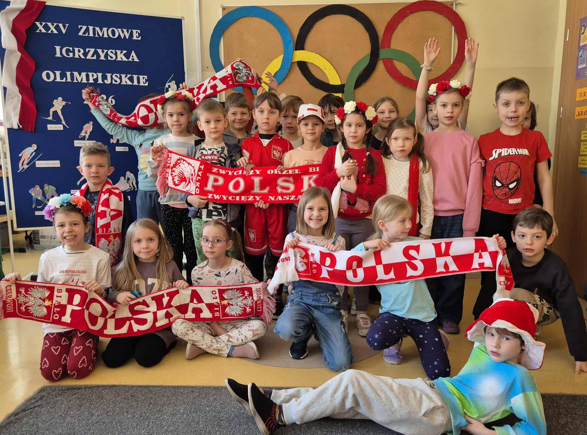 Grupa dzieci w sali przedszkolnej pozuje z biało-czerwonymi szalikami „Polska” na tle dekoracji z kołami olimpijskimi. Dzieci trzymają biało-czerwone szaliki z napisem „Polska” i mają na sobie elementy stroju w barwach narodowych. Obok znajduje się tablica informacyjna o Zimowych Igrzyskach Olimpijskich 2026.