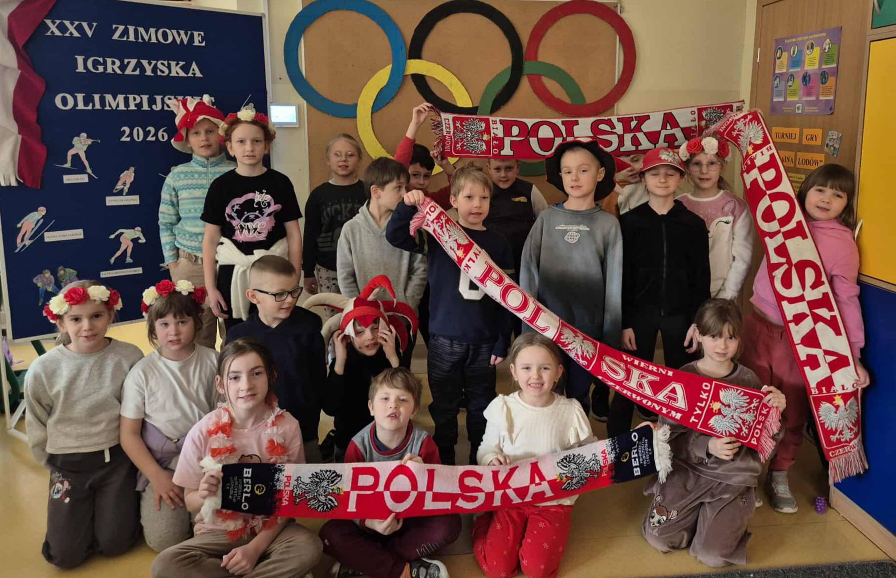 Grupa dzieci w sali przedszkolnej pozuje z biało-czerwonymi szalikami „Polska” na tle dekoracji z kołami olimpijskimi. Dzieci trzymają biało-czerwone szaliki z napisem „Polska” i mają na sobie elementy stroju w barwach narodowych. Obok znajduje się tablica informacyjna o Zimowych Igrzyskach Olimpijskich 2026.