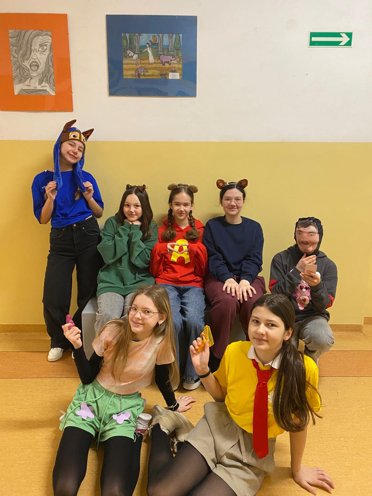 Grupa uczennic w przebraniach inspirowanych różnymi postaciami i stylami. Stoją i klęczą w szkolnym korytarzu, mają na sobie m.in. koszule, spódnice, pasy, chustę na głowie oraz kolorową perukę. W tle widoczne są prace plastyczne wywieszone na ścianie.