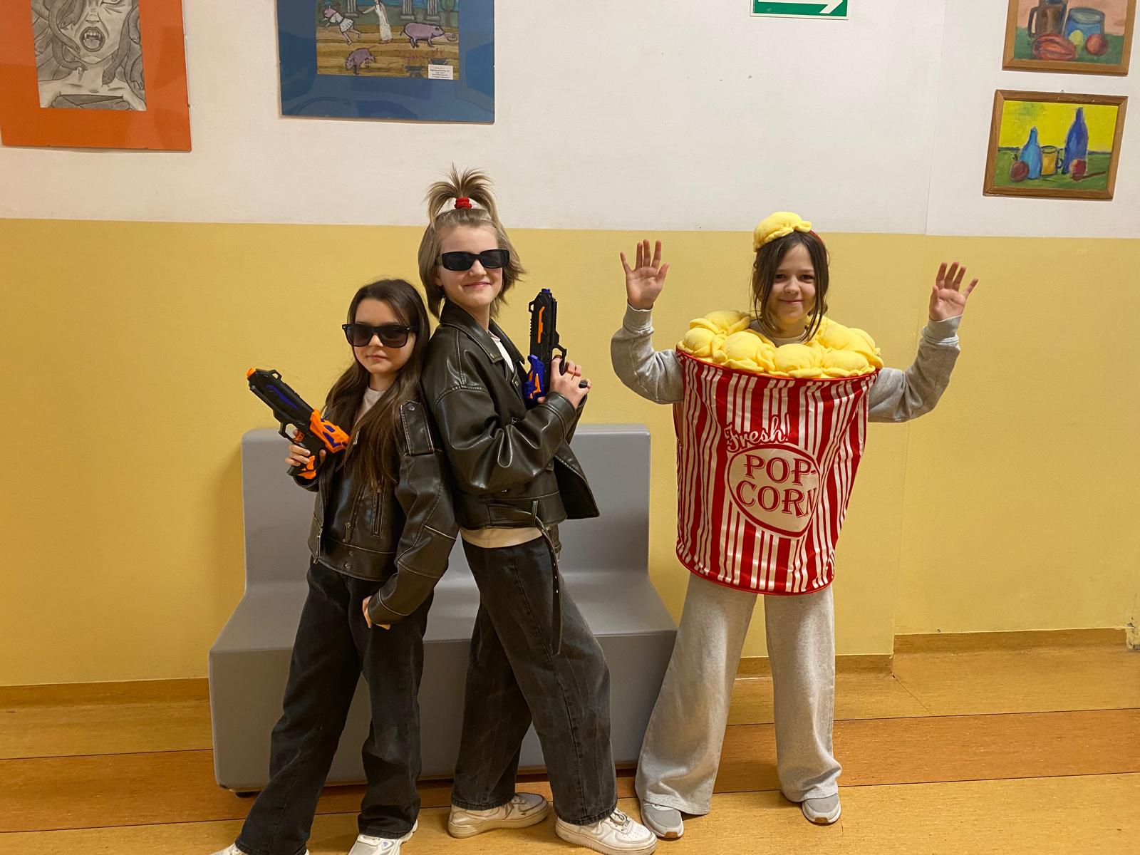 Trzy uczennice pozują w korytarzu szkolnym. Dwie z nich ubrane są w ciemne stroje z okularami przeciwsłonecznymi i trzymają zabawkowe pistolety, a trzecia ma zabawny kostium pudełka popcornu. Wszystkie uśmiechają się do zdjęcia.