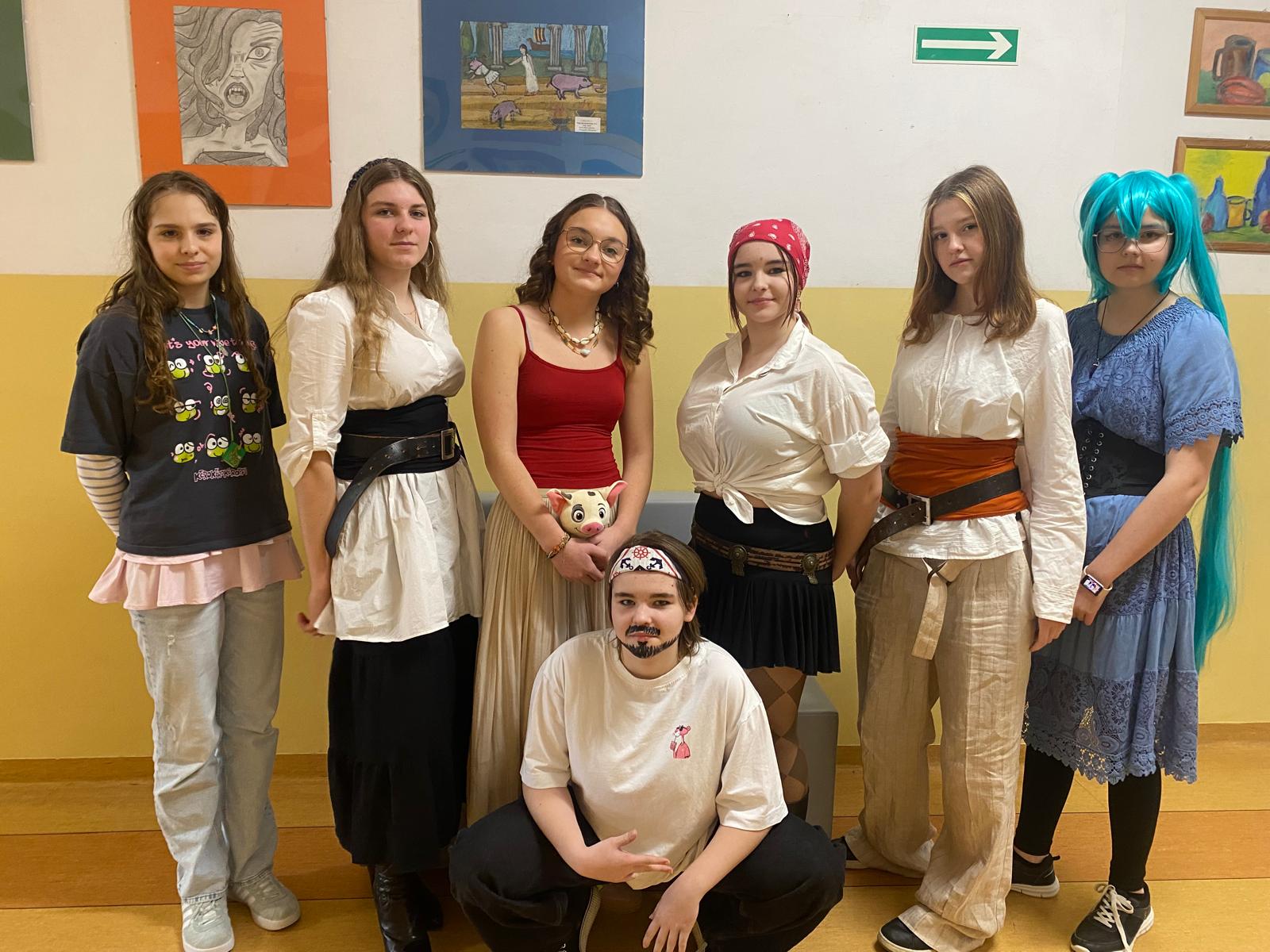 Grupa uczennic w przebraniach inspirowanych różnymi postaciami i stylami. Stoją i klęczą w szkolnym korytarzu, mają na sobie m.in. koszule, spódnice, pasy, chustę na głowie oraz kolorową perukę. W tle widoczne są prace plastyczne wywieszone na ścianie.