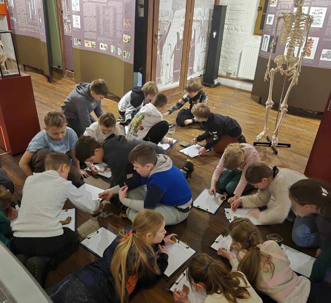 16 dzieci siedzi na podłodze. Dzieci rysują na kartkach swoje znaleziska .Obok dzieci stoi szkielet człowieka za dziećmi widać szyby w których są różne elementy muzeum w którym się znajdują .Są w muzeum archeologicznym.