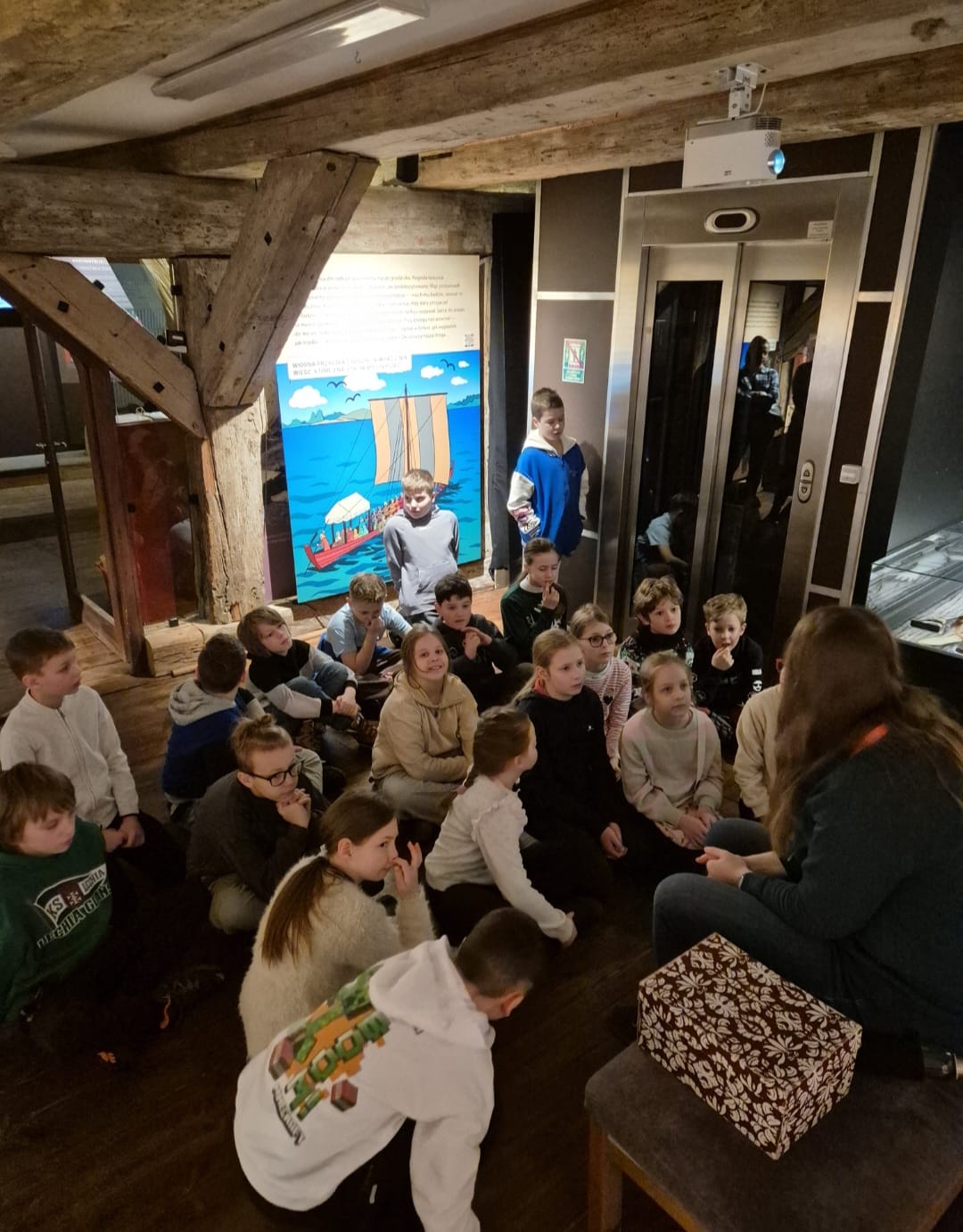 Grupa dzieci siedzi na podłodze i słucha Pani edukatorki. Jeden chłopiec stoi po prawej stronie sali, obok windy. Nad dziećmi znajduje się drewniany sufit. Obok Pani edukatorki  znajduje się pudełko. Za dziećmi znajduje się plakat z łódką w tle .Na jego tle widać chłopca który również stoi.