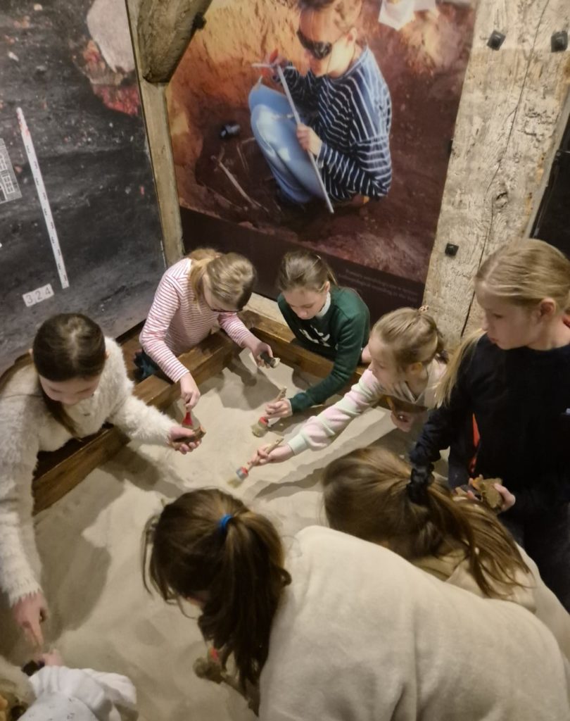 16 dzieci siedzi na podłodze. Dzieci rysują na kartkach swoje znaleziska .Obok dzieci stoi szkielet człowieka za dziećmi widać szyby w których są różne elementy muzeum w którym się znajdują .Są w muzeum archeologicznym.